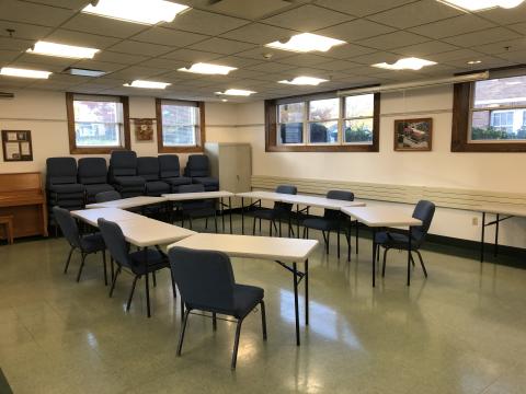 Image of Meeting Room 116 at Thorntown Public Library. Shows a set of tables and chairs with a piano, extra chairs, and storage in the background.